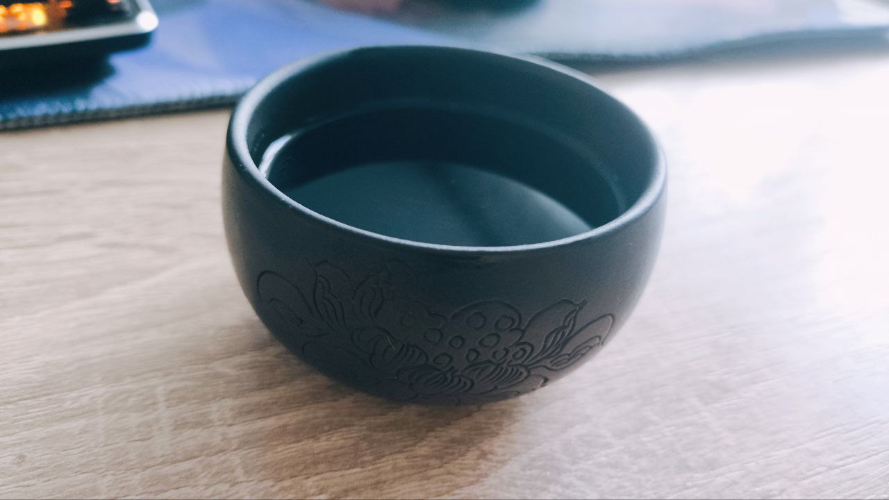 Black Ceramic Bowl on Wood A simple black ceramic bowl filled with clear liquid, sitting on a wooden surface.