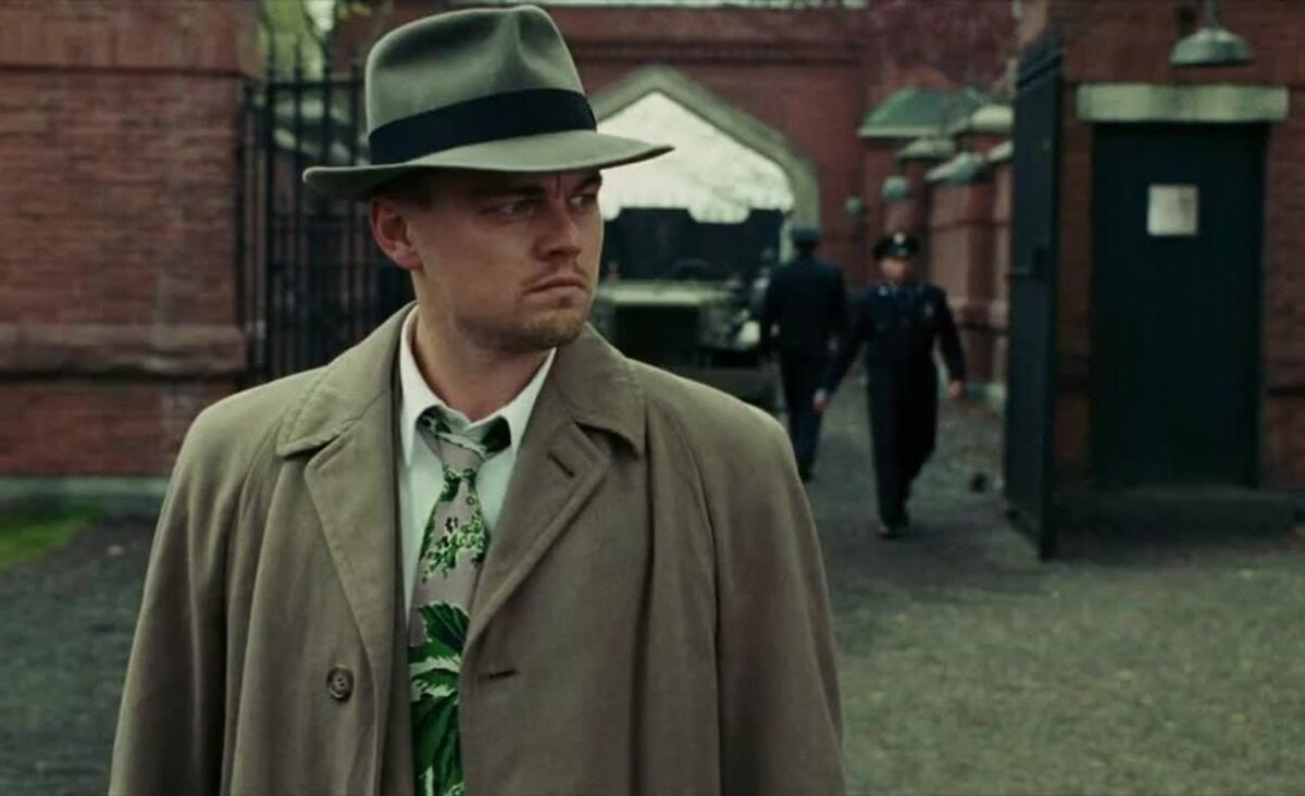 Man in Trench Coat with Thoughtful Gaze A man in a trench coat and fedora standing outdoors, looking intensely to the side, with a brick building and other figures in the background. His expression is serious.